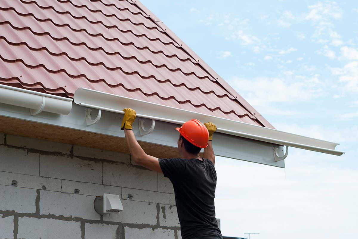 Installer in an orange hard hat fitting a white gutter onto the edge of a terracotta-tiled roof.