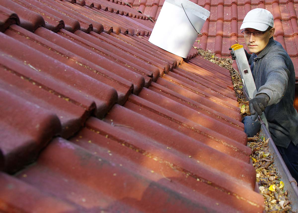 Gutter maintenance specialist clearing out debris from overflowing gutters on a red-tiled roof.